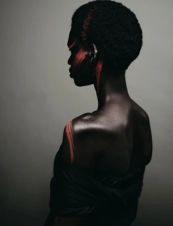 Profile portrait of a Black woman model with red face paint and hair accents against a neutral grey studio background.