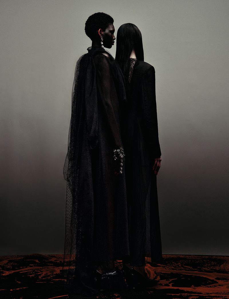 Two models in dark sheer garments standing back-to-back against a muted gradient background