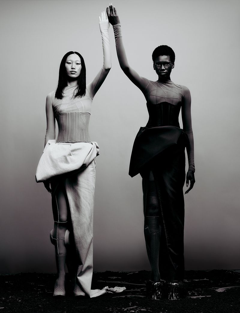 Two models standing side-by-side with raised hands touching, wearing structured avant-garde garments against a minimalist grey background.