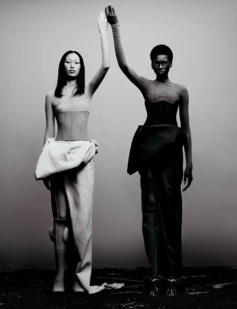 Two models standing side-by-side with raised hands touching, wearing structured avant-garde garments against a minimalist grey background.