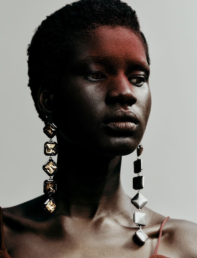 Close-up portrait of model Nyagoa Diu wearing long metallic statement earrings against a plain grey backdrop.