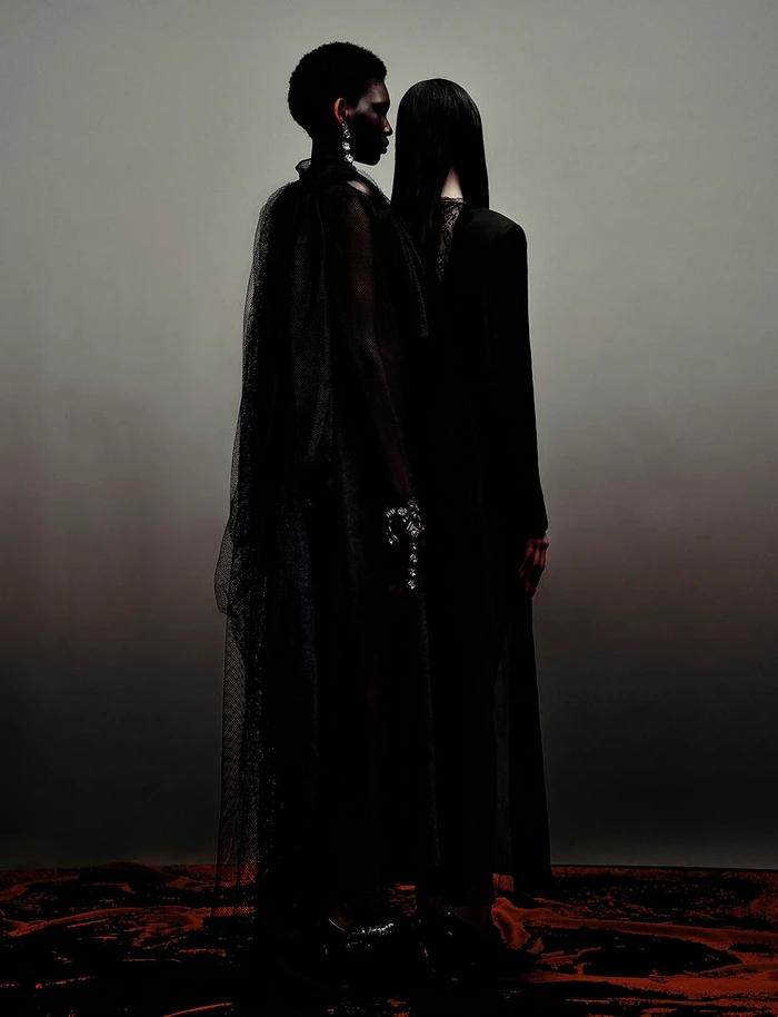 Two models in dark sheer garments standing back-to-back against a muted gradient background