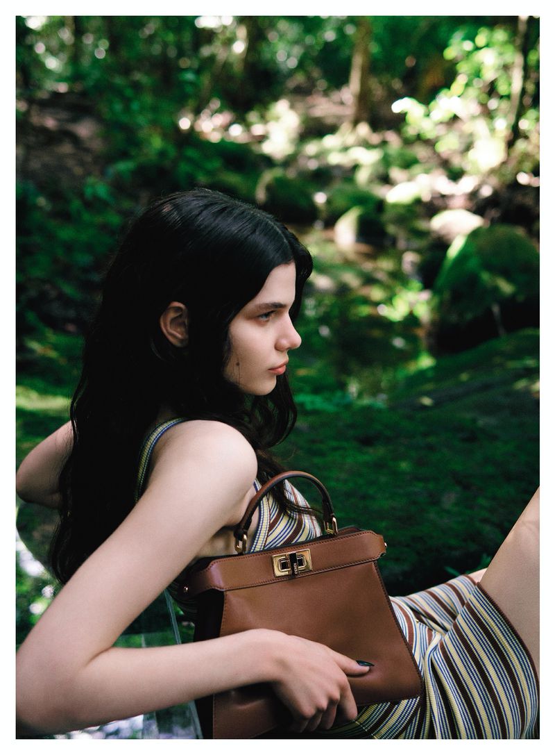 Woman with dark hair and a striped dress sits in a forest, holding a leather handbag, showcasing a fashion-forward scene in nature.