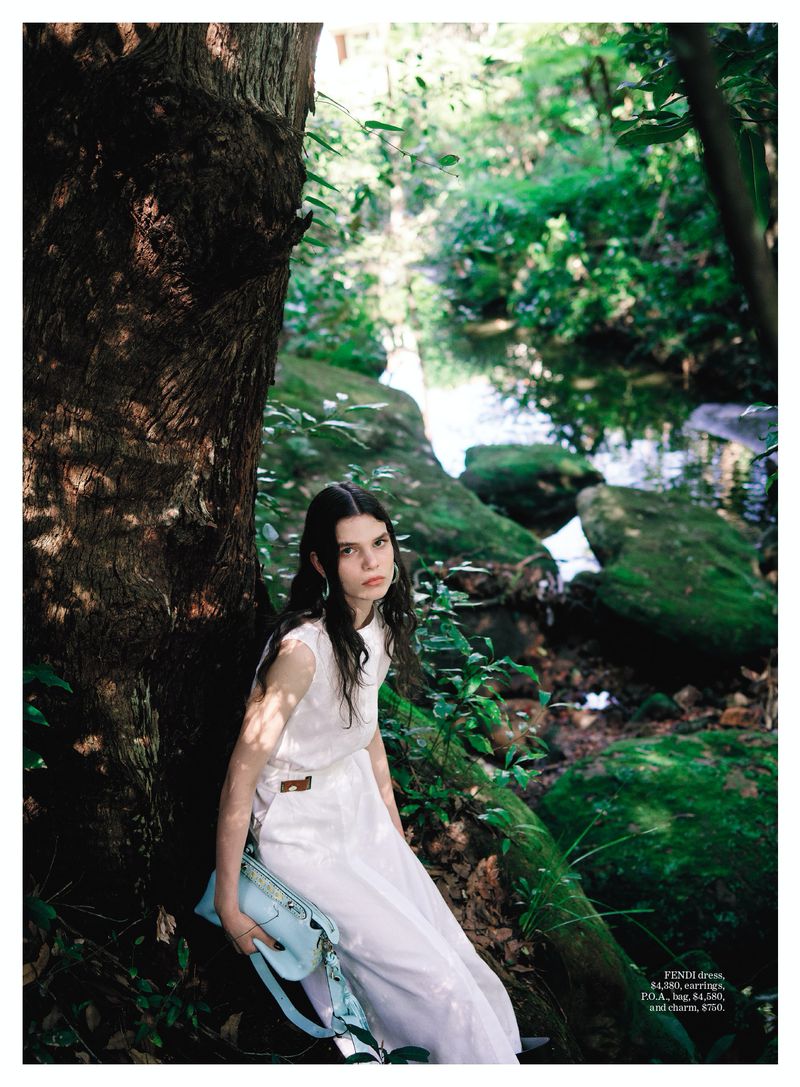 Model wearing a white dress leaning against a large tree trunk in a lush green forest with a stream