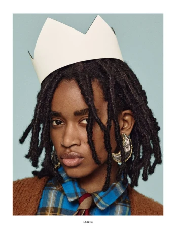 Close-up portrait of a person with dreadlocks wearing a paper crown, plaid shirt, tie, cardigan, and large earrings against a light blue background.