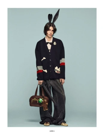 Model wearing a dark cardigan and wide-legged pants, accessorized with bunny ears and a brown satchel, against a light blue background.