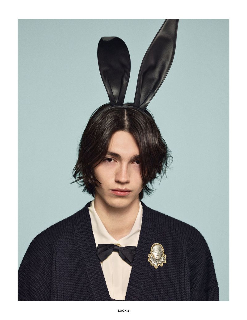 Portrait of a young adult male with long dark hair wearing rabbit ears, a black bow tie and a cardigan on a light blue background.