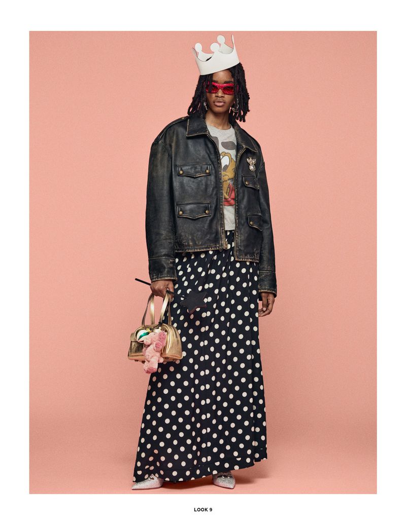 Fashion model poses in polka dot skirt, black jacket, and sunglasses with a small purse, wearing a crown in a studio setting.