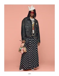 Model wearing a white paper crown, dark leather jacket, and polka dot skirt holding a handbag against a peach background.