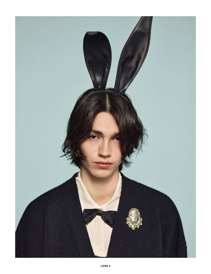 Model wearing black faux-leather bunny ears, a dark cardigan over a white shirt, a black bow tie, and a decorative brooch.
