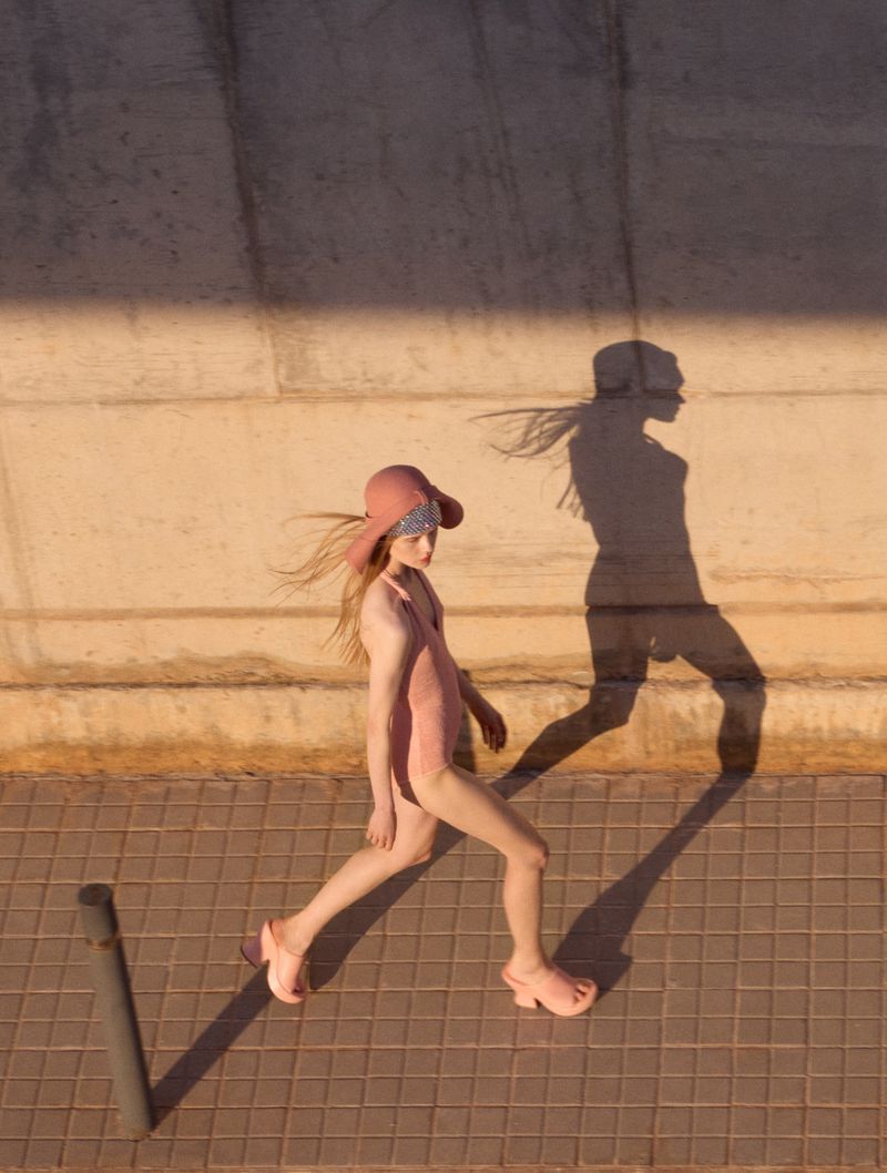 Young woman walks in retro bathing suit, sun hat, and platform sandals. Warm light casts her shadow on concrete wall.