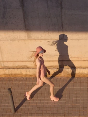 Young woman walks in retro bathing suit, sun hat, and platform sandals. Warm light casts her shadow on concrete wall.