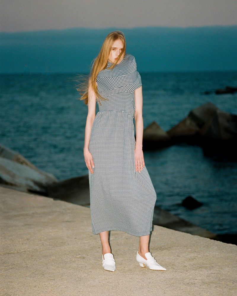 Model standing on a concrete pier in a patterned dress against an ocean and rocky coastline backdrop