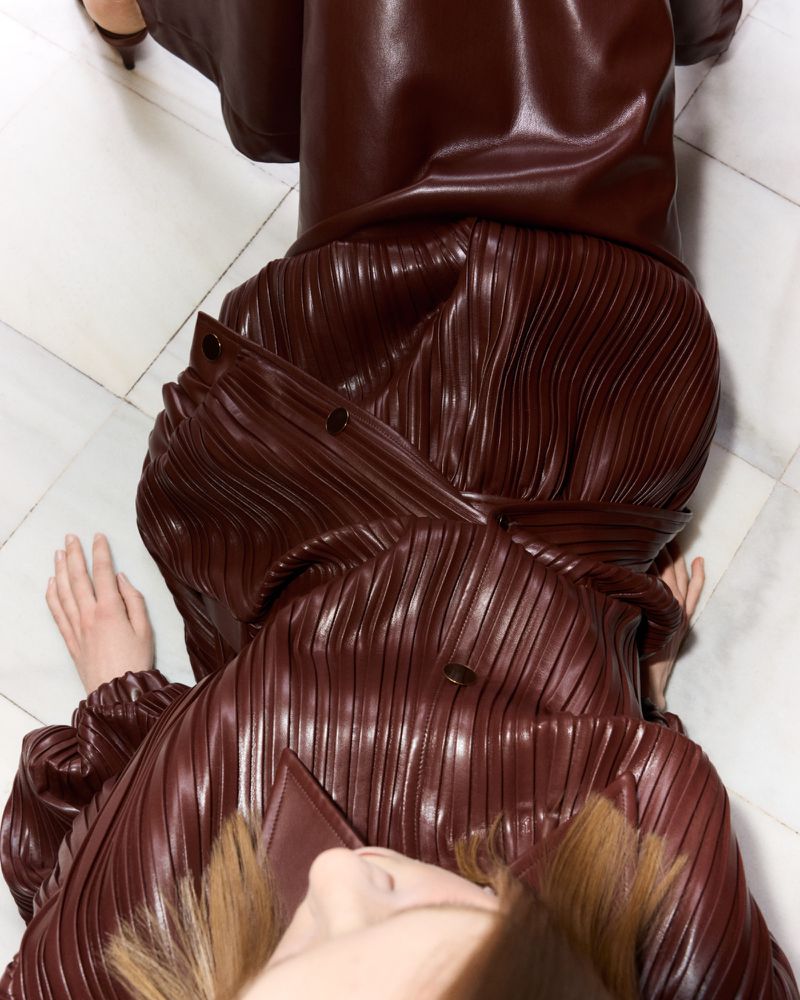 Model wearing dark brown pleated faux leather jacket reclining on white tiled floor