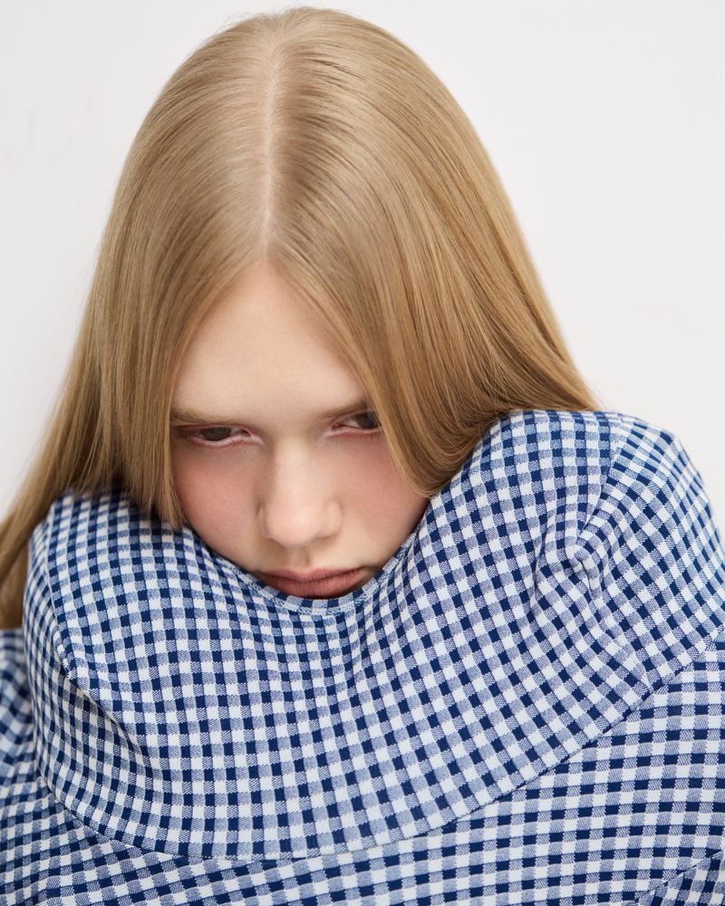 Close-up portrait of a model with blonde hair wearing a blue and white gingham patterned high-neck garment against a plain background.