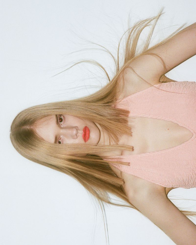 Model with long blonde hair and red lipstick wearing a pink textured top against a white background for Puss Puss Magazine.