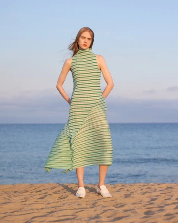 Model standing on a sandy beach wearing a green striped dress with the ocean and a clear sky behind.