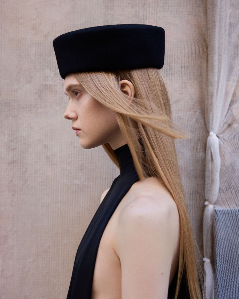 Profile portrait of a blonde woman wearing a black pillbox hat and black high-neck top for Puss Puss Magazine Barceloneta project