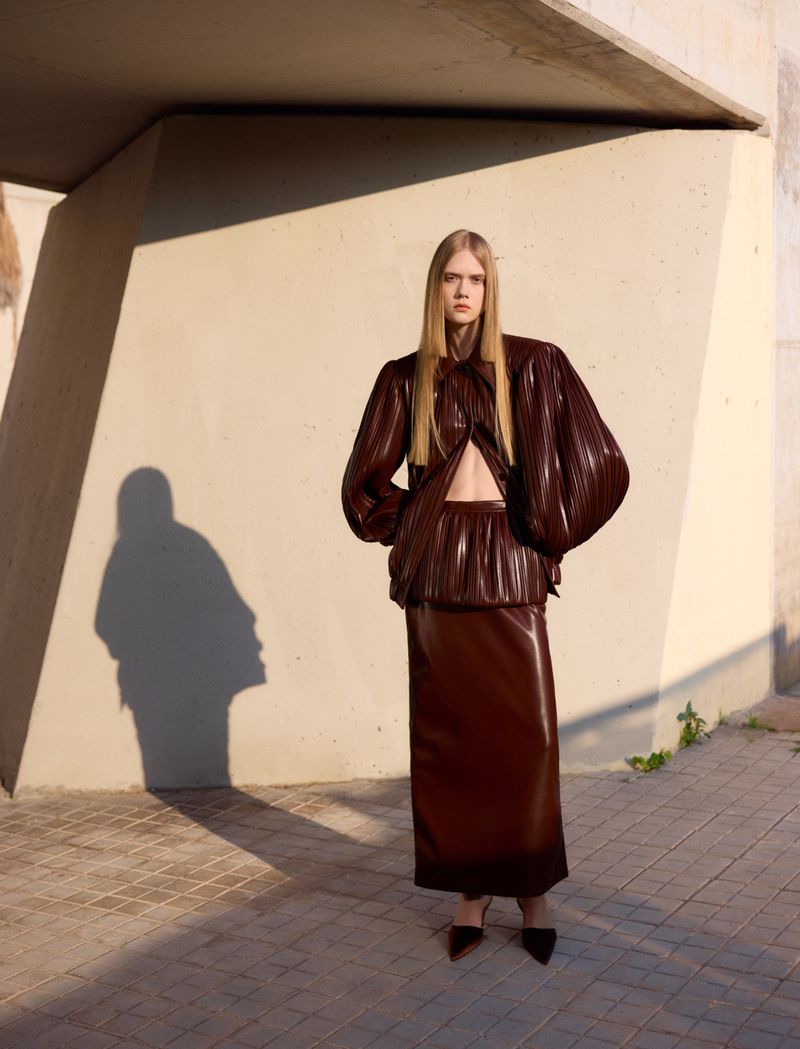 Model standing outdoors wearing a shiny brown pleated two-piece outfit against a concrete architectural wall with long shadows.