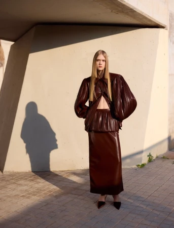 Model standing outdoors wearing a shiny brown pleated two-piece outfit against a concrete architectural wall with long shadows.