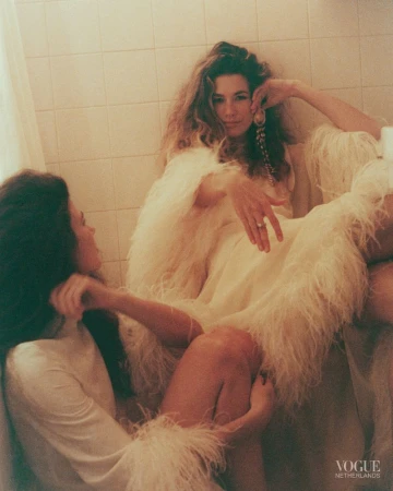 Two women in feathered and white dresses posing in a tiled bathroom for an editorial shoot by Vogue Netherlands