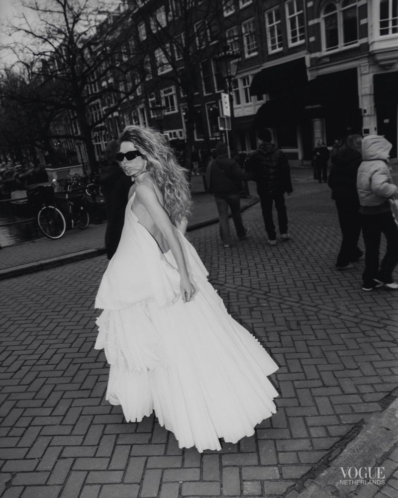Model in white tiered dress and sunglasses walking on a cobblestone street with buildings and pedestrians in background.