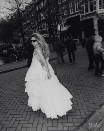 Model in white tiered dress and sunglasses walking on a cobblestone street with buildings and pedestrians in background.