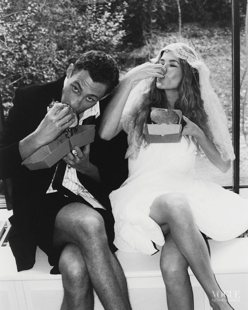 Black and white fashion editorial image of a man and a bride eating fast food burgers inside