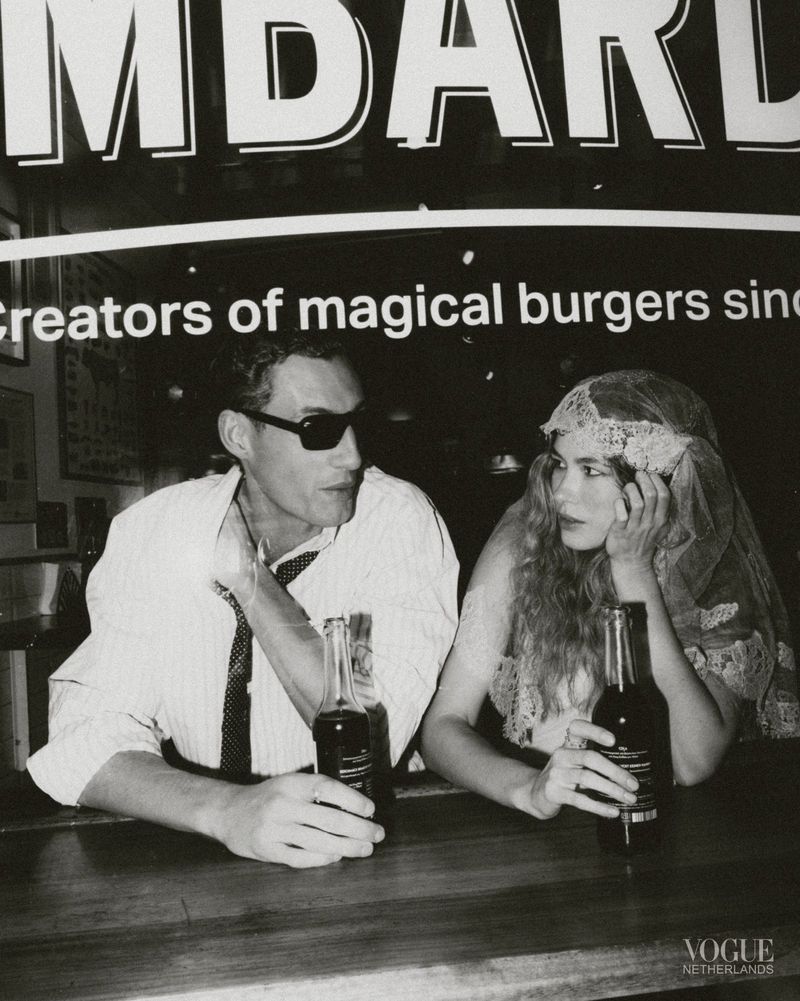 Black and white editorial image of a man and a woman seated at a counter holding glass bottles beneath a large sign.