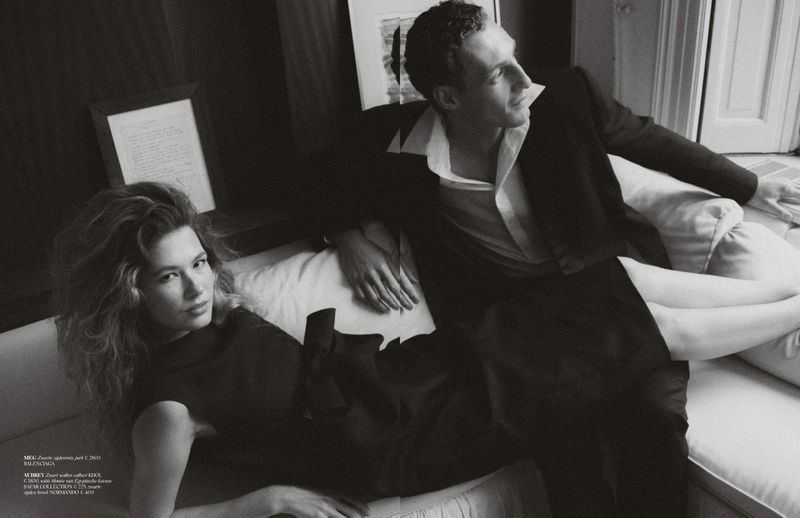 Black and white editorial portrait of a woman reclining and a man seated on a light couch in a room.