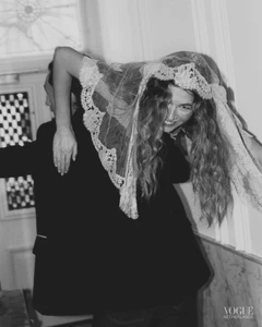 Black and white editorial image of a smiling woman wearing a lace veil draped over her head, being carried by a man in a dark suit.