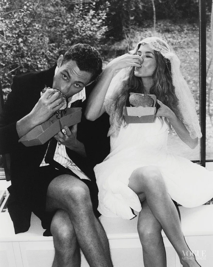 Black and white fashion editorial image of a man and a bride eating fast food burgers inside