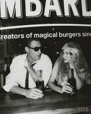 Black and white editorial image of a man and a woman seated at a counter holding glass bottles beneath a large sign.