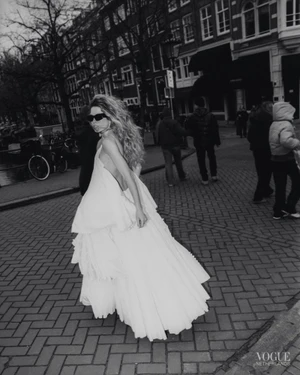 Model in white tiered dress and sunglasses walking on a cobblestone street with buildings and pedestrians in background.
