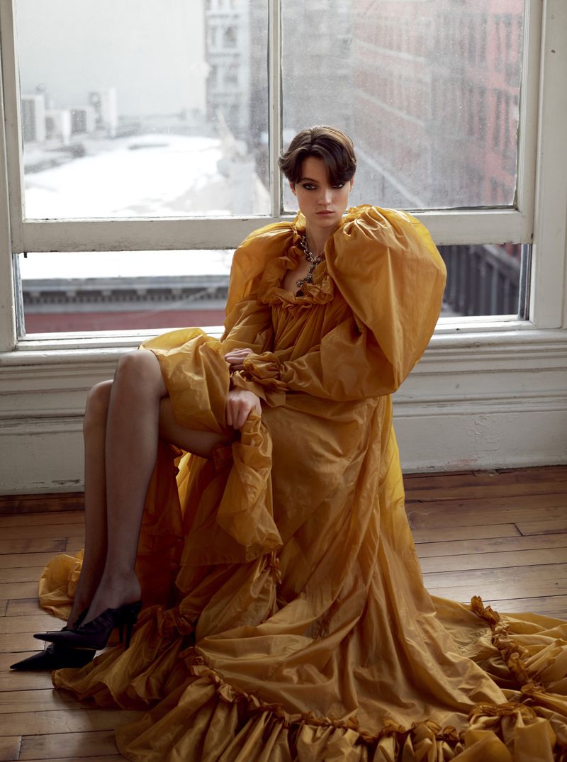 Model seated in a voluminous gold ruffled dress by a large window overlooking a city street.