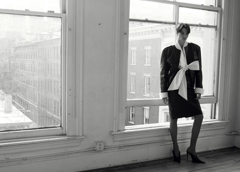 Model standing by large windows wearing a dark jacket with a white bow tie for HERO Magazine