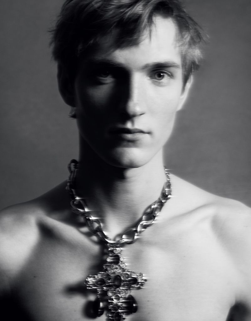 Portrait of a man wearing a large ornate cross pendant necklace against a plain dark background.
