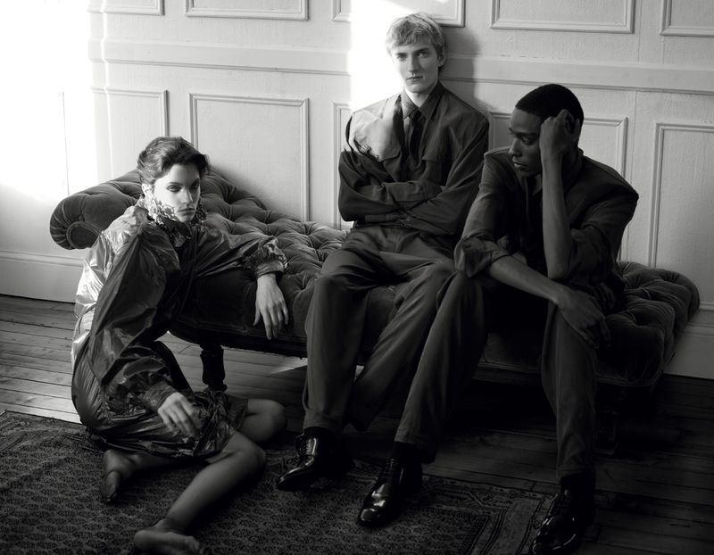 Monochrome fashion editorial image showing three models posing on and around a tufted chaise lounge indoors.