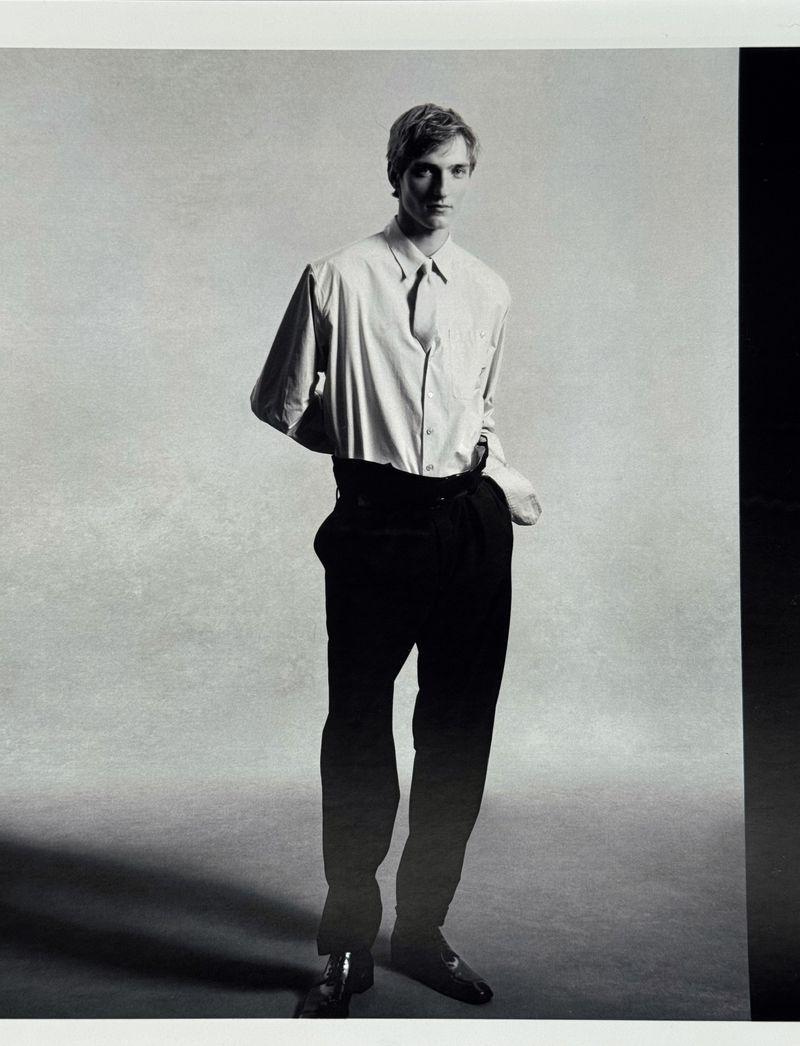 Full-length portrait of a male model wearing a light-coloured collared shirt and dark trousers against a neutral background.