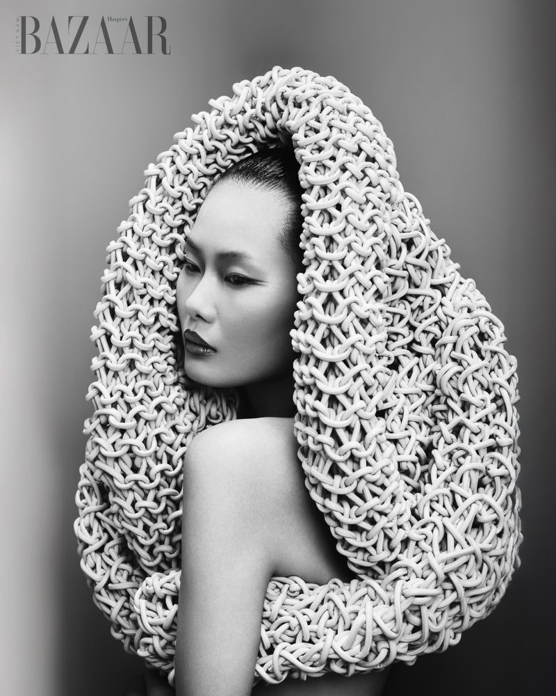 Asian woman framed inside a large sculptural knitted headpiece in a black and white editorial portrait for Harper's Bazaar Vietnam.