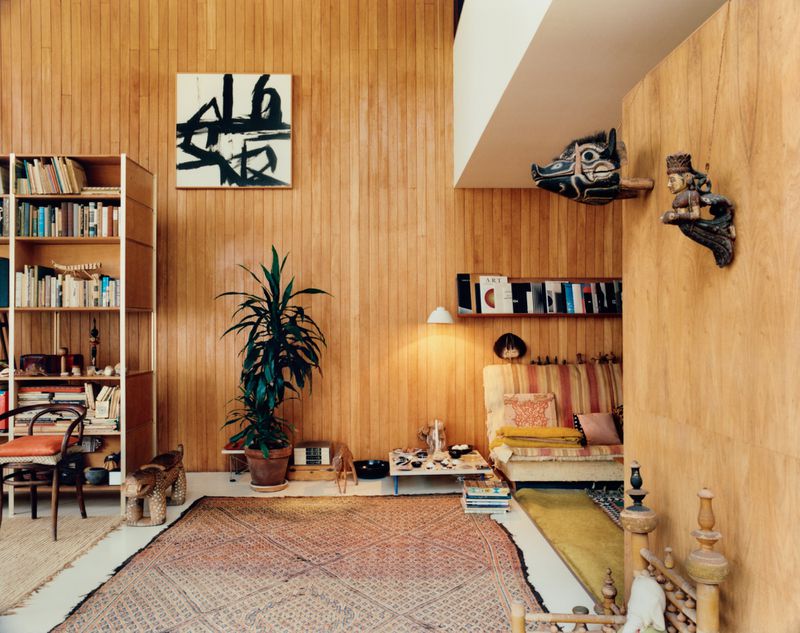Interior view of the Eames House featuring warm wood paneling, bookshelves, a potted plant, and patterned rug.