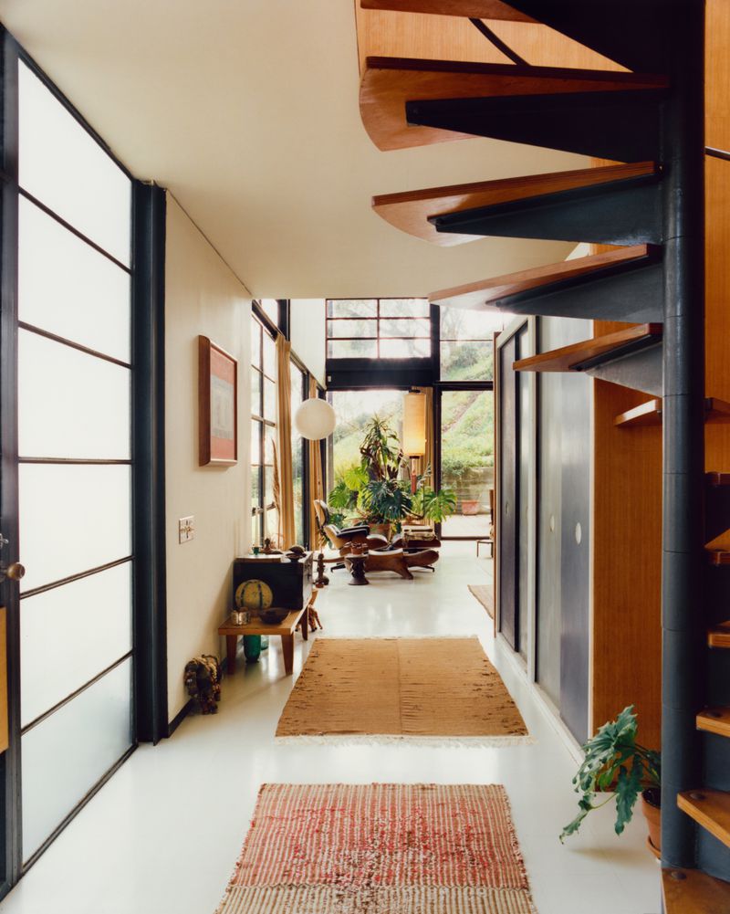 Interior view of the Eames House featuring a prominent wooden spiral staircase and a long hallway with rugs on a white floor.
