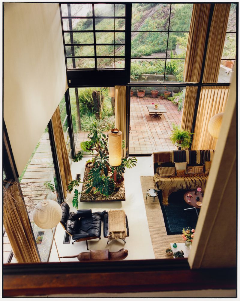 High-angle interior view of the Eames House living room featuring large glass windows, indoor plants, and mid-century modern furniture.