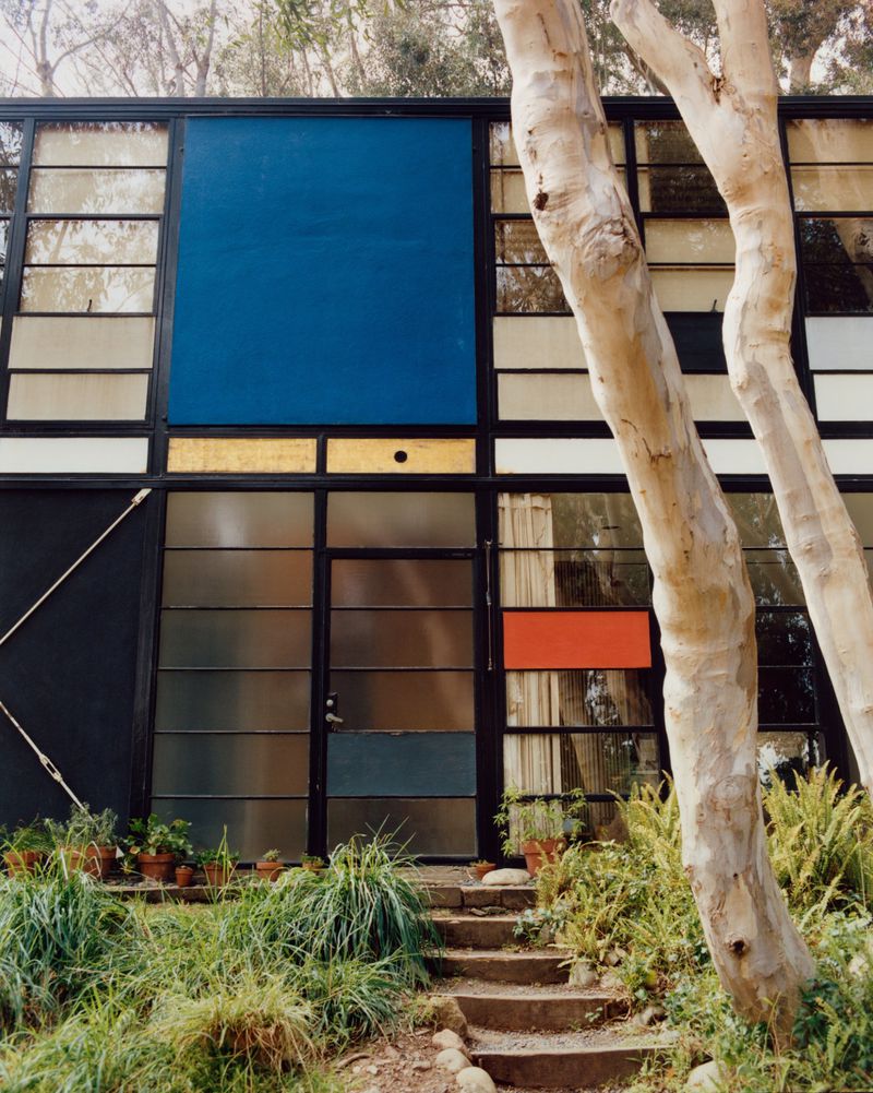 The Eames House exterior showing steel frame architecture with blue and orange panels, framed by eucalyptus trees and foliage.