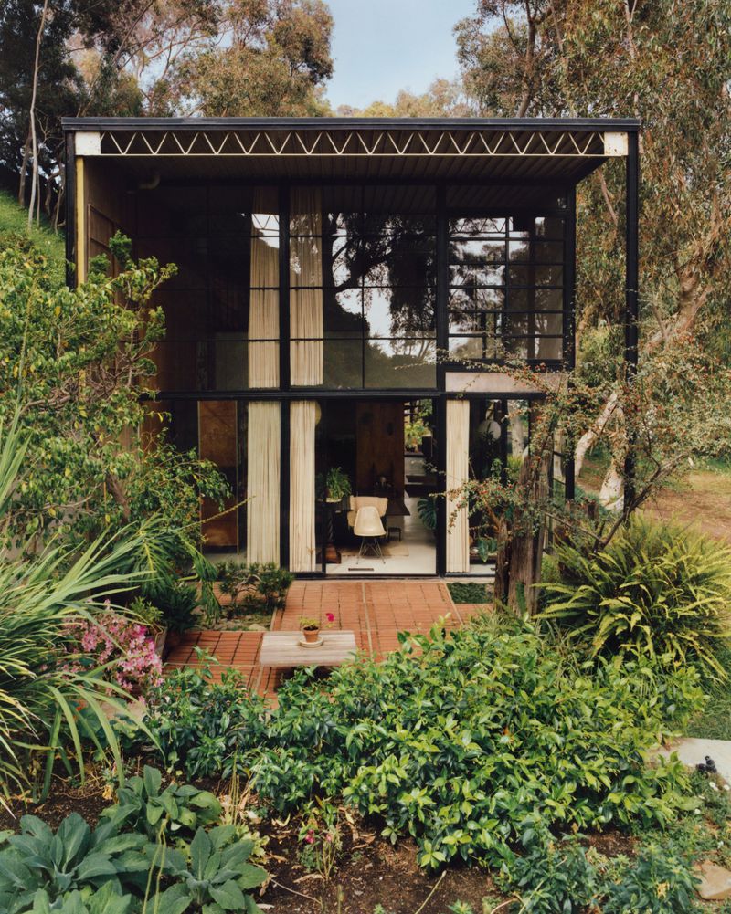 Exterior view of The Eames House surrounded by lush green trees and foliage with large glass facade and steel structural framing.