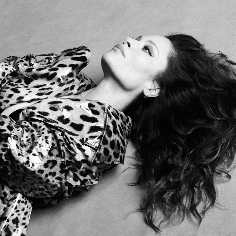 Portrait of a woman with long brown hair, wearing a leopard print top, looking up in a black and white studio shot with a light background.