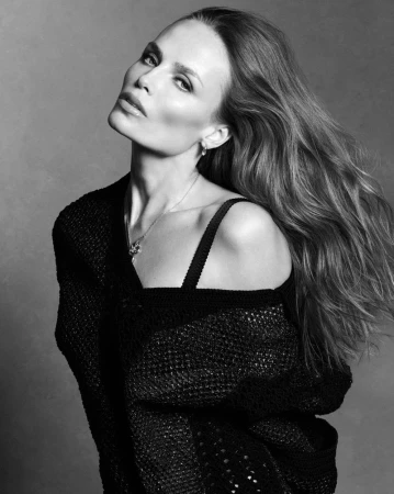 Model Natasha Poly with flowing long hair wearing a black knitted top against a grey textured studio backdrop.