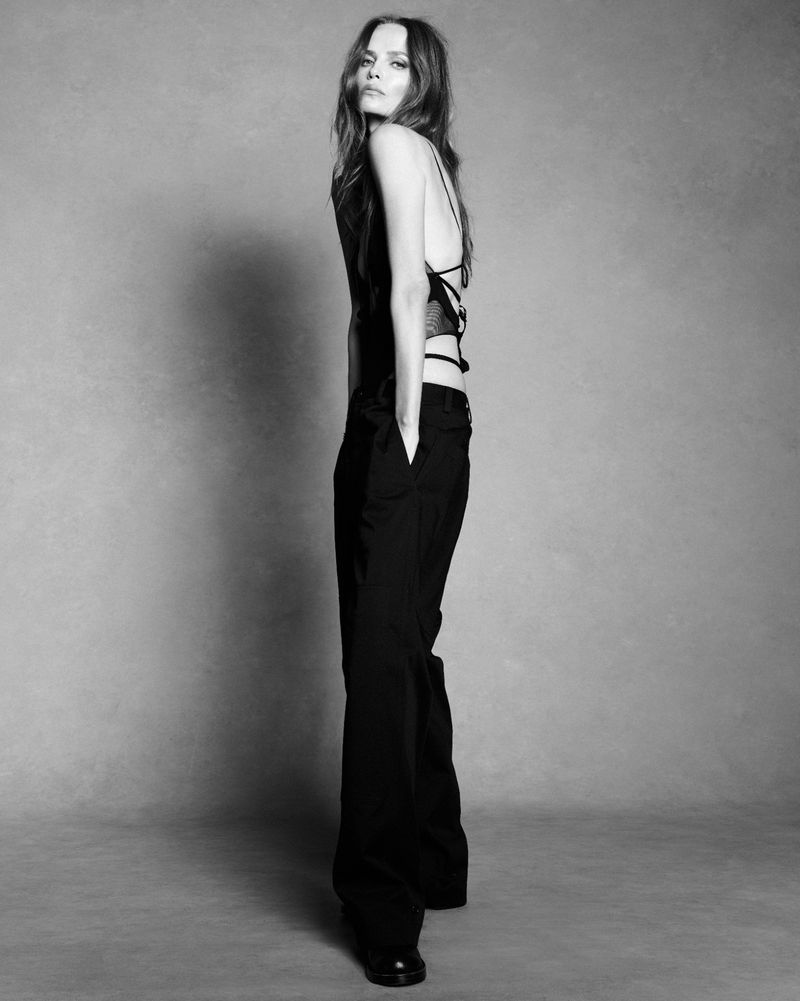 Model Natasha Poly standing in profile wearing dark trousers and a black top against a seamless grey studio backdrop