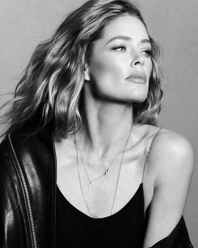 Monochrome portrait of model Doutzen Kroes wearing a dark jacket and layered necklaces against a grey studio backdrop.