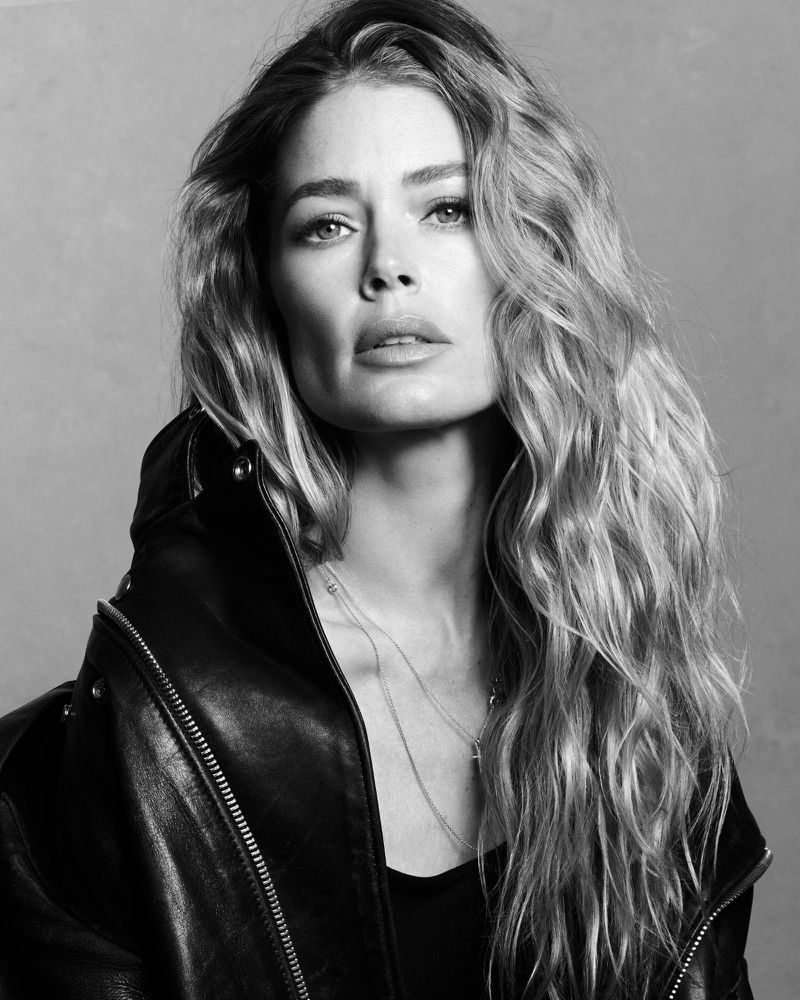 Monochrome portrait of model Doutzen Kroes wearing a leather jacket and layered necklaces against a textured grey background.